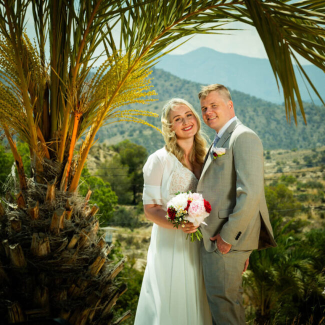 Wedding portrait of the bride and groom smiling at each other, dressed in wedding attire, with a natural outdoor background.